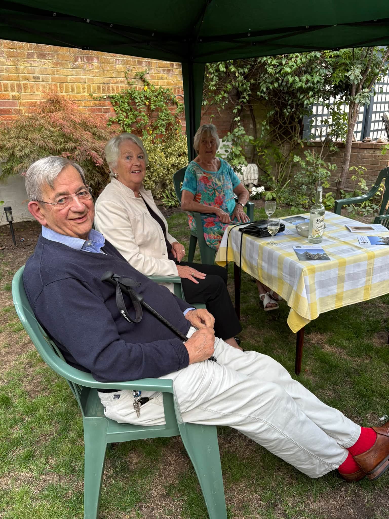 2026 Friends Summer Party and AGM 3 people sitting round a small table under a gazebo. There are drinks of wine and juice on the table